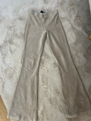Beige mjukisbyxor med utsvängda ben - Supersköna beige mjukisbyxor i mjukt velourmaterial med utsvängda ben. Perfekta för en avslappnad och trendig look. Hög midja och stretchig passform gör dem riktigt bekväma att bära hela dagen.