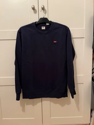Mörkblå sweatshirt från Levi's - Mörkblå sweatshirt från Levi's med rund halsringning och liten röd logotyp på bröstet. Tröjan har långa ärmar och ribbade muddar vid ärmslut och nederkant. Perfekt för en avslappnad och stilren look.