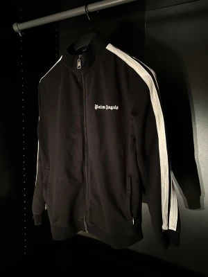 Svart track jacket från Palm Angels - Svart track jacket från Palm Angels med vita ränder längs ärmarna och dragkedja framtill. Jackan har hög krage, ribbade muddar och logga broderad på bröstet. Perfekt för en sportig och trendig look.