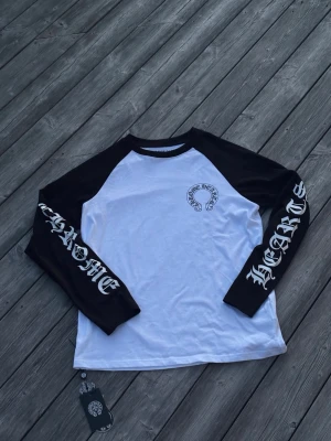 Chrome Hearts långärmad t-shirt - Säljer en svartvit långärmad t-shirt från Chrome Hearts med tryck på bröstet och stora logotexter längs båda ärmarna. Klassisk baseballmodell med svarta ärmar och vit kropp. Perfekt för dig som gillar streetwear och vill sticka ut.