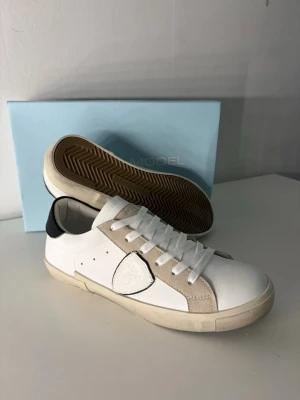 Vita Philippe Model sneakers med beige detaljer - Snygga vita sneakers från Philippe Model med beige mockadetaljer och svart hälparti. Skorna har klassisk låg profil, vita skosnören och gummisula med fiskbensmönster undertill. Perfekta för dig som gillar stilrena och trendiga sneakers.
