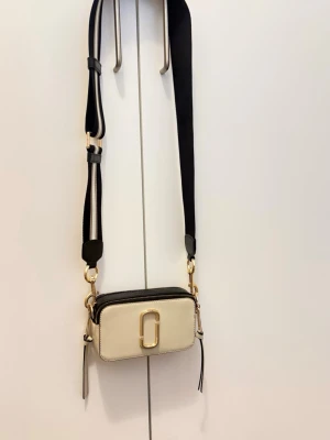 Marc Jacobs Snapshot axelväska beige - Snygg beige och svart axelväska från Marc Jacobs med bred justerbar axelrem och guldfärgade detaljer. Väskan har två fack med dragkedja och ikonisk logga framtill. Tillverkad i tåligt läder och canvas. Perfekt för dig som gillar trendiga accessoarer.