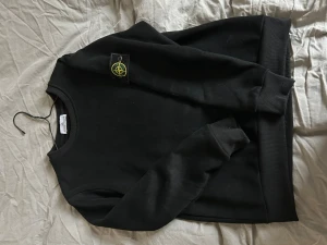 Svart sweatshirt från Stone Island - Säljer en svart sweatshirt från Stone Island med den klassiska loggan på vänster ärm. Tröjan har rund halsringning, ribbade muddar vid ärmslut och nederkant samt en avslappnad passform. Perfekt för en stilren och enkel look.