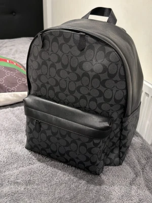 Svart ryggsäck från Coach - Snygg svart ryggsäck från Coach med det klassiska monogrammönstret över hela väskan. Tillverkad i slitstarkt konstläder med en stor ytterficka framtill och dragkedja. Perfekt för dig som vill ha en stilren och trendig look.