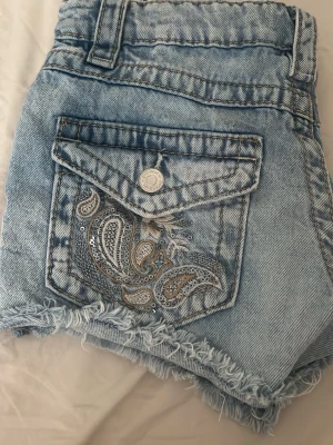 Blå denimshorts med paljettdetalj - Säljer ett par ljusblå denimshorts från Gina  med hög midja och fransig kant. Baksidan har en ficka med paisleybroderi och silverpaljetter som ger extra bling. Perfekta för sommaren och riktigt snygga till sneakers eller sandaler. Har använt bara 2 gånger inga fläckar !