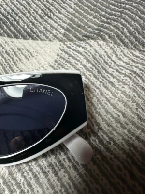 Svarta och vita Chanel solglasögon - Säljer ett par riktigt snygga solglasögon från Chanel med svart båge och vita skalmar. Glasen är mörka och bågarna har en cool, futuristisk form som verkligen sticker ut. Perfekta för dig som vill ha en statement-accessoar från ett lyxigt märke.