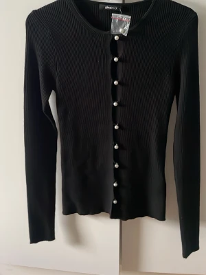 Svart ribbad kofta från Gina Tricot - Snygg svart ribbad kofta från Gina Tricot med långa ärmar och rund halsringning. Koftan har dekorativa pärlknappar framtill och är aldrig använd.