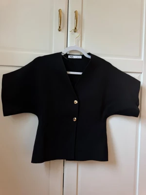 Svart kortärmad topp från ZARA - Stilren svart topp från ZARA med v-ringning och två guldfärgade knappar framtill. Modellen har breda, korta ärmar och en rak, något boxig passform. Perfekt att styla till både jeans och kjol för en trendig look.