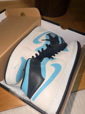 Nike Jordan - Snygga Nike Air Jordan 1 Mid sneakers i vitt, ljusblått och svart läder. Klassisk siluett med svart snörning, blå swoosh och Air Jordan-logga på sidan. Perfekta för dig som vill sticka ut med stilrena och trendiga sneakers.