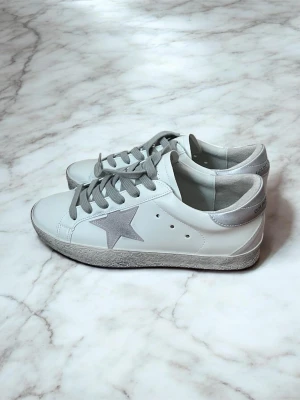 Golden Goose silvertail modellen - Snygga vita sneakers från Golden Goose med grå snörning, silverfärgad stjärna på sidan och metallic-detaljer på hälen. Skorna har en klassisk låg profil och är tillverkade i skinn för en exklusiv känsla.