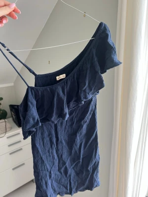 Marinblå omlottklänning Hollister - Söt marinblå blus från Hollister med omlottdesign och volangdetalj längs halsringningen. Blusen har smala axelband och är tillverkad i ett lätt, strukturerat tyg som passar perfekt till varmare dagar.