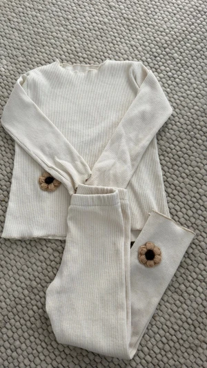 Beige ribbat loungewear-set med blommor - Mysigt loungewear-set i beige ribbat tyg med långärmad tröja och matchande byxor. Setet har dekorativa virkade blommor i beige och brunt på både tröjan och byxbenet. Perfekt för chill dagar hemma.