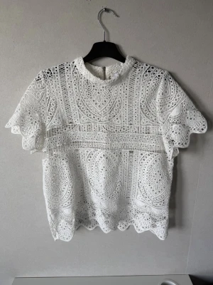 Vit spetsblus med kort ärm - Supersnygg vit blus i spets med korta ärmar och hög hals. Blusen har ett detaljerat broderat mönster och vågig kant nertill. Dragkedja bak för enkel påklädning. Blusen är helt ny men utan prislapp. 