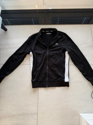 Svart ziptröja från Lacoste - Snygg svart ziptröja från Lacoste med vita paneler på sidorna och klassisk krokodillogga på bröstet. Tröjan har hög krage, hel dragkedja och ribbade muddar. Perfekt för en sportig och clean look.
