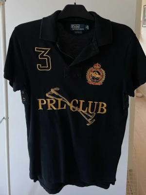 Svart Polo Ralph Lauren  piké /t-shirt - Svart t-shirt/ piké från Polo Ralph Lauren med gul broderad text 'PRL CLUB', poloklubba och nummer 3 på bröstet. Märkets emblem i rött och guld på vänster sida. Klassisk pikékrage och korta ärmar. Perfekt för dig som gillar sportig och stilren look.