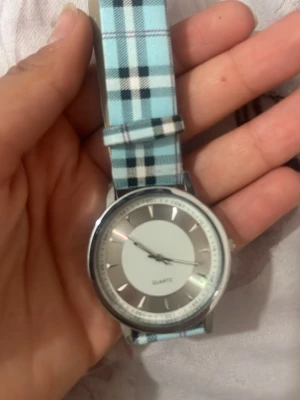 Blårutig klocka med silverfärgad urtavla - Snygg klocka med blårandigt och rutigt band i textil och en stilren silverfärgad urtavla. Klockan har minimalistisk design och passar perfekt för dig som gillar en fräsch och modern accessoar. Quartz-urverk och klassisk rund form.