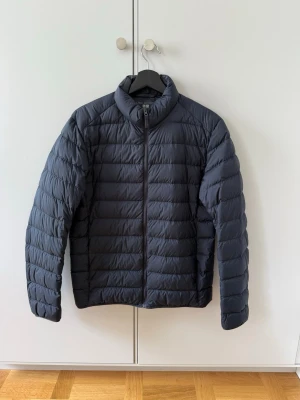 Mörkblå pufferjacka  - Snygg mörkblå pufferjacka från Uniqlo med hög krage och hel dragkedja framtill. Jackan har quiltad design med horisontella sömmar och långa ärmar. 