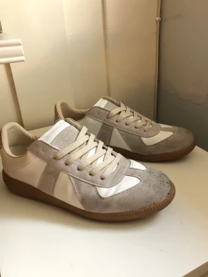 Vita och beige sneakers med mockadetaljer - Säljer ett par stilrena sneakers i vitt och beige med klassiska mockadetaljer och gummisula. Skorna har låga skaft, vita snören och en retroinspirerad design som passar perfekt till jeans eller cargo. Materialet är en mix av mocka och slätt läder.