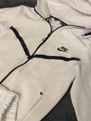 Vita mjukisbyxor från Nike - Snygga vita mjukisbyxor från Nike med svarta detaljer och dragsko i midjan. Byxorna har elastisk midja, sidofickor och Nike-logga på låret. Perfekta för en chill och sportig look.