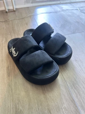 Svarta platåsandaler Juicy Couture - Säljer ett par svarta platåsandaler från Juicy Couture med breda band, sandalerna är i mocka  och har en stor glittrig logga i metall på sidorna av båda sandalerna. Snygg chunky sula och riktigt statement-vibe. Perfekta för dig som vill sticka ut. Använda vid endast 2 tillfällen. 