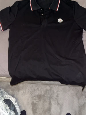 Svart Moncler piké - Snygg svart pikétröja från Moncler med klassisk krage och knappar. Kragen och ärmsluten har röda och vita ränder som ger en clean kontrast. Moncler-logga på bröstet. Perfekt till jeans eller shorts för en stilren look.