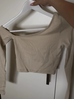 Beige croppad långärmad topp - Trendig beige croppad topp med lång ärm och bred, något asymmetrisk halsringning. Perfekt att styla med höga jeans eller kjol för en clean look. Mjuk och stretchig känsla, passar dig som gillar minimalistiskt mode.