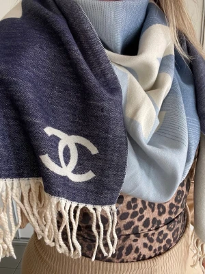 Blå och vit halsduk från Chanell - Snygg halsduk från Chanell i blått och vitt med klassisk logga. Halsduken har fransar i ändarna och är tillverkad i ett mjukt, vävt material som känns lyxigt och skönt runt halsen. Perfekt accessoar för att lyfta din outfit.