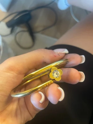 Guldigt armband med blomdetalj - Snyggt armband i guldfärgad metall med en unik form och en blomformad detalj framtill. I mitten av blomman sitter en glittrande sten som ger extra bling. Perfekt accessoar för dig som vill sticka ut med något annorlunda och trendigt.