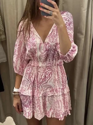 Rosa och vit kortklänning med volang - Söt kortklänning i rosa och vitt med blommigt mönster, v-ringning och trekvartsärm. Klänningen har volangdetaljer och resår i midjan för en bekväm passform. Perfekt för sommarens alla tillfällen.