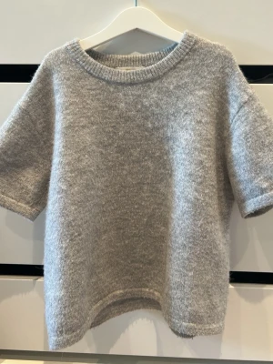 Grå stickad tröja Gina Tricot - Mjuk och fluffig grå stickad tröja från Gina Tricot med korta ärmar och rund halsringning. Tröjan har ribbstickade detaljer vid hals, ärmslut och nederkant. Perfekt för lager på lager och enkel att matcha med jeans eller kjol. Tröjan är ganska noprig,men inget man tänker på när man väl använder den.