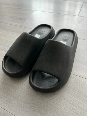 Svarta slides från Adidas - Säljer ett par svarta slides från Adidas med öppen tå och bred rem över foten. Tillverkade i mjukt och lätt syntetmaterial för maximal komfort. Perfekta att glida runt i hemma eller på stranden. Klassisk och stilren design med Adidas-logga på sulan.
