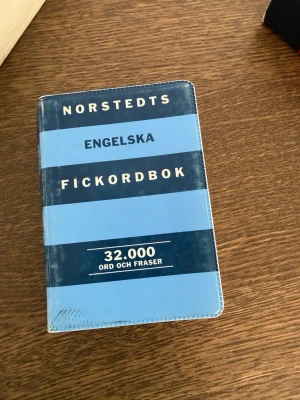 Norstedts Engelska Fickordbok - Praktisk fickordbok i blått och svart med 32 000 engelska ord och fraser. Perfekt att ha med sig i väskan eller fickan när du pluggar eller reser. Mjukpärm och smidig storlek gör den enkel att använda när som helst.