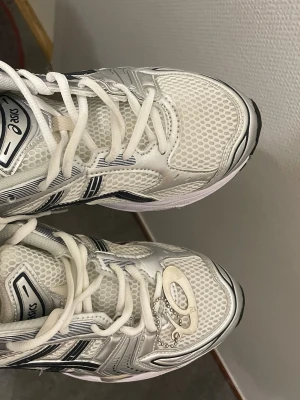 Vita Asics sneakers med mesh och silver - Säljer ett par vita Asics sneakers med mesh-överdel och silverdetaljer. Skorna har svarta och grå inslag samt klassisk Asics-logga på sidan. Låga i modellen och med vita snören. Perfekta för dig som gillar sportig och clean stil.