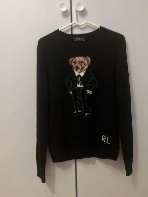 Svart stickad tröja Polo Bear RL - Stickad svart tröja från Polo Ralph Lauren med den ikoniska Polo Bear i mitten, klädd i mörka kläder. RL-broderi nere vid fållen. Rund halsringning och långa ärmar. Perfekt för dig som gillar klassiska streetwear-vibbar. Storlek xs (160-168) pris kan diskuteras vid snabb affär 