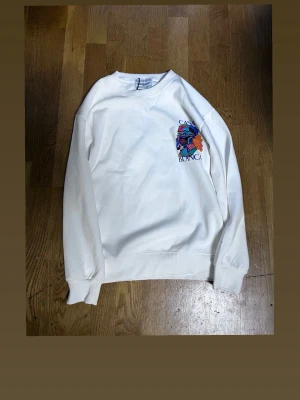 Vit sweatshirt från Casablanca - Säljer en vit sweatshirt från Casablanca med färgglatt tryck och texten 'CASABLANCA' på bröstet. Tröjan har rund halsringning, långa ärmar och ribbade muddar. Perfekt för dig som gillar streetwear och unika prints. OBS LITEN I STLRK!!!!!