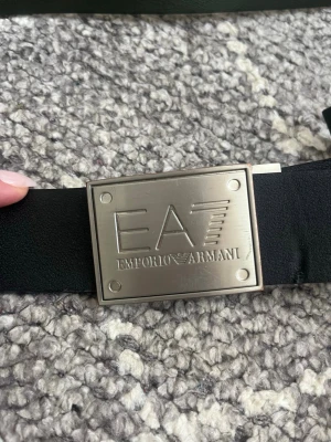 Svart skärp från Emporio Armani - Snyggt svart bälte från Emporio Armani med en stor, silverfärgad metallspänne där loggan EA är ingraverad. Bältet har en klassisk och stilren design som passar till många olika outfits. Perfekt accessoar för dig som gillar exklusiva detaljer. 112 cm. Lite missfärgningar på metallen 