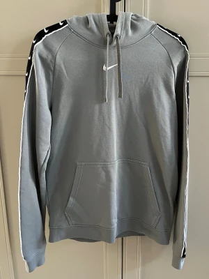 Grå Nike hoodie med logga på ärmar - Snygg grå hoodie från Nike med huva och dragsnören. Svarta ränder med vita Nike-loggor längs båda ärmarna. Känguruficka framtill. Mjukt material.