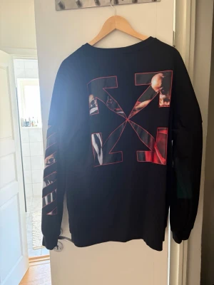 Svart sweatshirt från Off-White - Säljer en svart sweatshirt från Off-White med stora grafiska tryck på ryggen och ärmen samt logga och handmotiv på bröstet. Tröjan har rund hals och långa ärmar. Materialet är mjukt bomullstyg och passformen är relaxed fit. Perfekt för dig som gillar streetwear och unika prints.