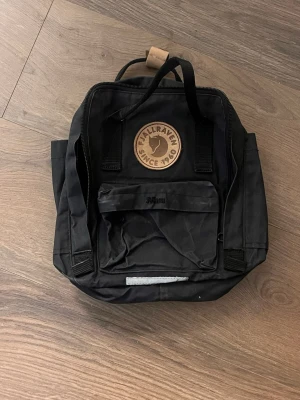 Svart Mini Kånken ryggsäck Fjällräven - Säljer en svart Mini Kånken ryggsäck från Fjällräven med det klassiska runda lädermärket framtill. Ryggsäcken har justerbara axelband, bärhandtag upptill och en liten framficka med dragkedja. Tillverkad i slitstarkt material och har sidofickor.
