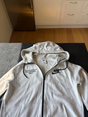 Vit Nike hoodie med tryck - Snygg vit hoodie från Nike med svart tryck 'AUTHORIZED PERSONNEL ONLY' på bröstet och ryggen. Klassisk Nike-logga på framsidan. Dragkedja framtill och stor huva. Perfekt för dig som gillar streetwear och vill sticka ut lite extra.