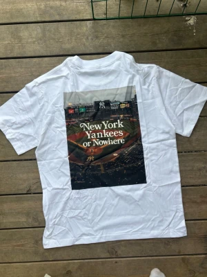 Vit New York Yankees t-shirt - Snygg vit t-shirt från New York Yankees med stort tryck på ryggen av en basebollarena och texten 'New York Yankees or Nowhere'. På bröstet finns en liten basebollhandske med boll. Perfekt för dig som gillar sportig streetstyle.