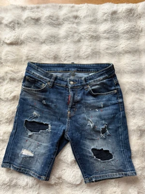 Blå slitna jeansshorts från Dsquared2 - Säljer ett par blå jeansshorts med slitna detaljer. Köpta på Garms Market. Bra skick och perfekt för sommaren. Lappen baktill är något sliten/lite lossnad, men det påverkar varken passform eller användning.