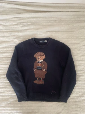 Mörkblå Polo Bear stickad tröja - Stickad tröja i mörkblått från Polo Ralph Lauren med ikoniska Polo Bear-motivet framtill. Rund halsringning och ribbade muddar vid ärmslut och nederkant. Tröjan har RL-broderi nedtill och är gjord i mjukt material, perfekt för en klassisk och cool look.