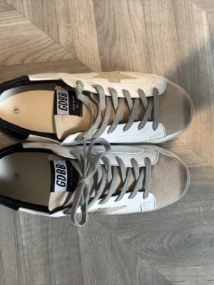  vita och beige sneakers med stjärna - Snygga sneakers från i vitt läder med beige mockadetaljer och grå snörning. Skorna har en  stjärna på sidan och svart hälparti. Perfekta för en trendig och avslappnad stil. Det är INTE golden goose!!    Skorna kommer med en i en vit box 
