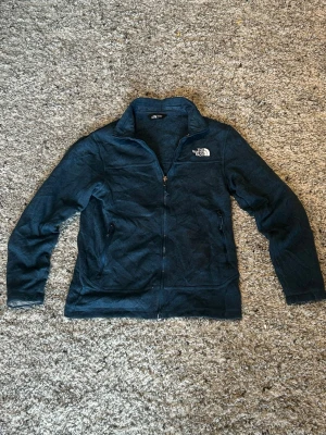 Mörkblå fleecejacka The North Face - Mysig mörkblå fleecejacka från The North Face med dragkedja framtill och två sidofickor med dragkedja. Jackan har hög krage och broderad logga på bröstet. Perfekt för lager-på-lager och chill dagar.