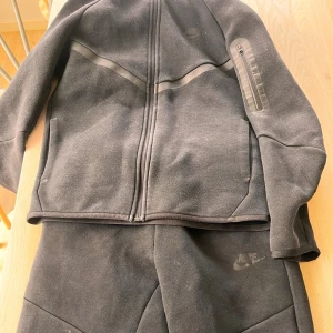 Svart Nike loungewear set - Mysigt svart loungewear set från Nike med långärmad ziptröja och matchande byxor. Tröjan har dragkedja framtill och en ficka med dragkedja på ärmen. Perfekt för chill dagar hemma eller när du vill vara bekväm. Tröjan är 147 158 och byxorna är en storlek mindre.