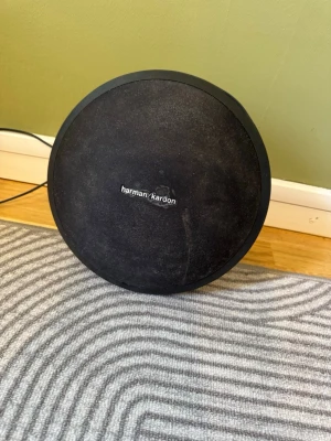 Harman Kardon Onyx Studio högtalare - Snygg trådlös högtalare från Harman Kardon i svart med rund form och stilren design. Högtalaren har bärhandtag, metallfötter och tygfront. Perfekt för att streama musik via Bluetooth och ger riktigt bra ljudkvalitet. Obs! Batteriet är slut så man måste ha strömsladden som följer med i när man använder de’