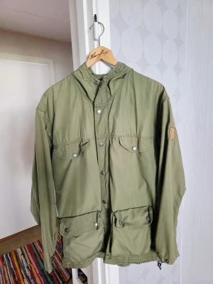 Grön vindjacka från Fjällräven - Snygg grön vindjacka från Fjällräven med huva, stora fickor framtill och klassisk logotyp på ärmen. Jackan har tryckknappar och dragsko nertill. Perfekt för uteliv och äventyr, tillverkad i slitstarkt material.