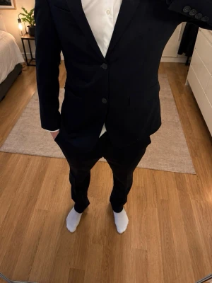 Svart kostym med kavaj och byxor från dressman - Stilren svart kostym med klassisk kavaj och matchande byxor. Kavajen har två knappar framtill och en smal krage. Perfekt för dig som vill ha en tidlös och snygg look. Passar till skjorta och slips för en uppklädd stil.