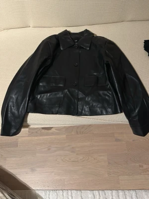 Svart skinnjacka med fickor - Snygg svart skinnjacka med klassisk krage och stora framfickor. Jackan har rak passform och stängs med knappar framtill. Perfekt för dig som vill ha en stilren och tidlös look.